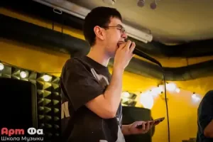 Совсем юный парень вытягивает ноту, микрофон прижат к губам, в другой руке телефон с текстом – Квартирник 07.06, июнь 2026